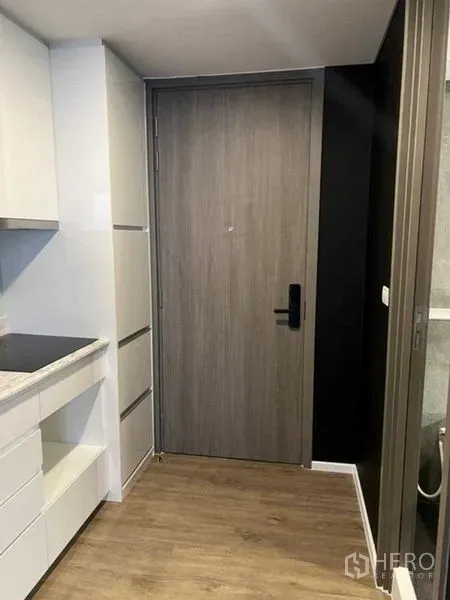 Condominium for sale in Huai Khwang, Bangkok - Entryway with wood door and compact kitchenette featuring white cabinets and cooktop.
