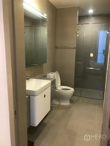 Condominium for rent in Pathum Wan, Bangkok - Modern bathroom featuring a vanity with mirrored cabinet, toilet, and glass-enclosed shower in neutral tiles.