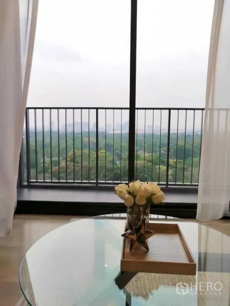 Condominium for rent in Chatuchak, Bangkok - Balcony with floor-to-ceiling glass and railing overlooking lush park views, with a vase of flowers on the coffee table.