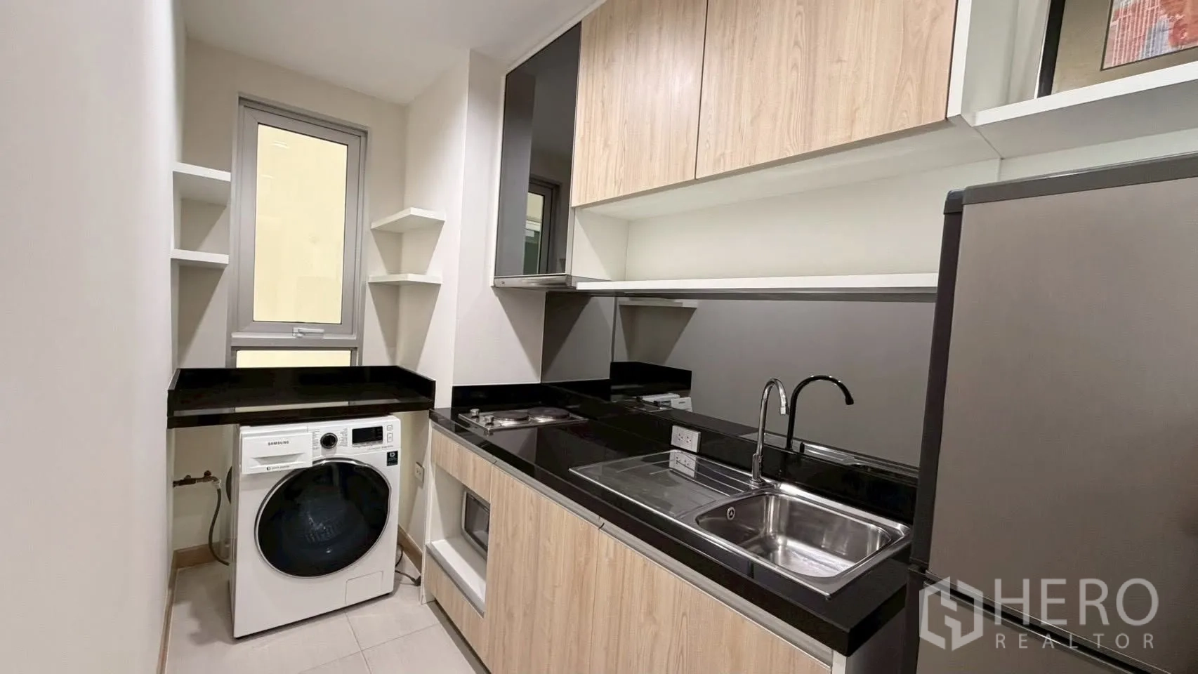 Condominium for rent or sale in Watthana, Bangkok - Separate galley kitchen with black countertops, sink, cooktop, cabinets, refrigerator, and washing machine.