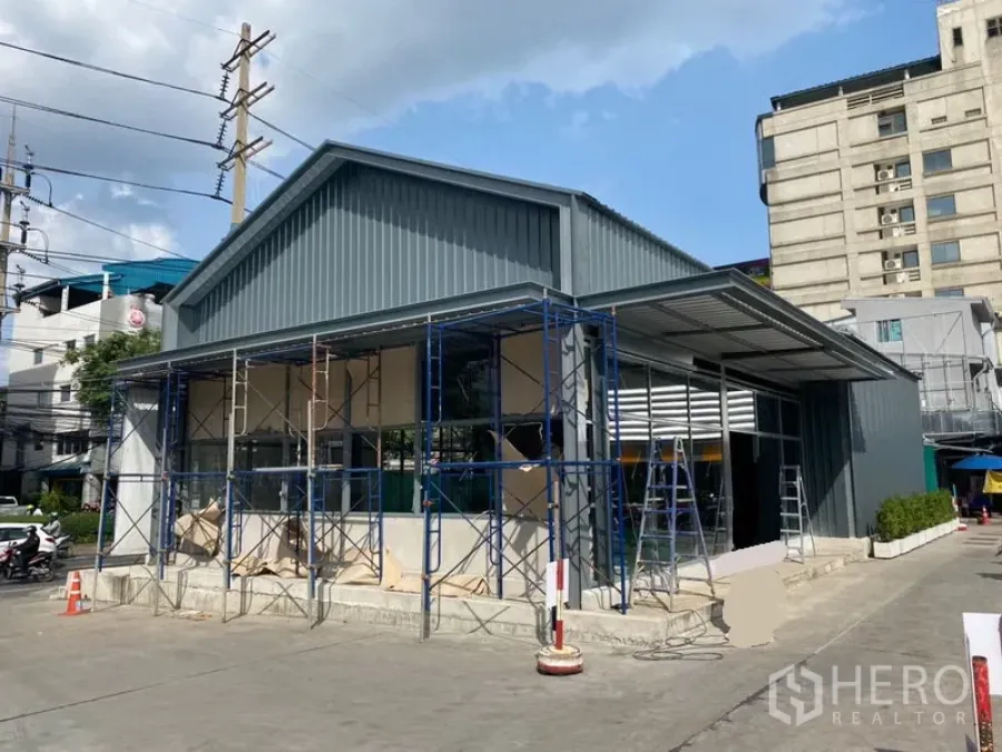 零售空间 出租在Khlong Toei, Bangkok - 单层现代零售建筑，玻璃立面与金属外墙，施工脚手架可见，前方设有停车空地，位于空堤区城市街景中。