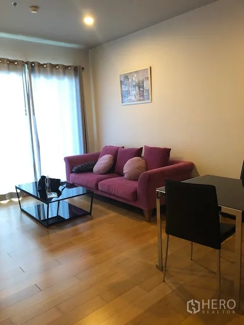 公寓 出租在Khlong San, Bangkok - 明亮客厅，紫色沙发、抱枕、玻璃茶几与落地窗帘。