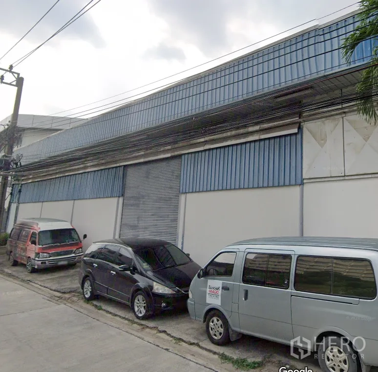 Warehouse for rent in Suan Luang, Bangkok - Street view of an industrial warehouse facade with blue cladding, large roller shutter door and parking in front.
