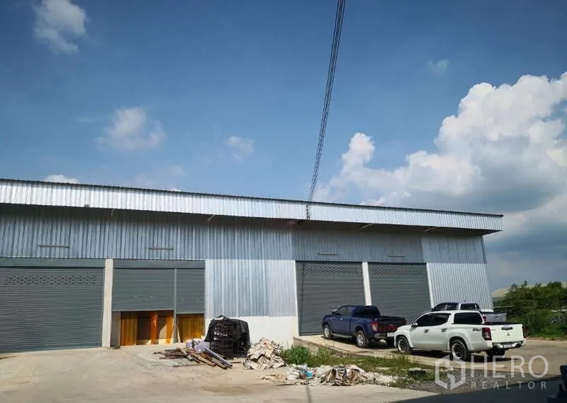 货栈 出租在Khlong Luang, Pathum Thani - 金属外墙仓库外观，灰色卷闸门，前方停车位与皮卡车，蓝天下的园区。