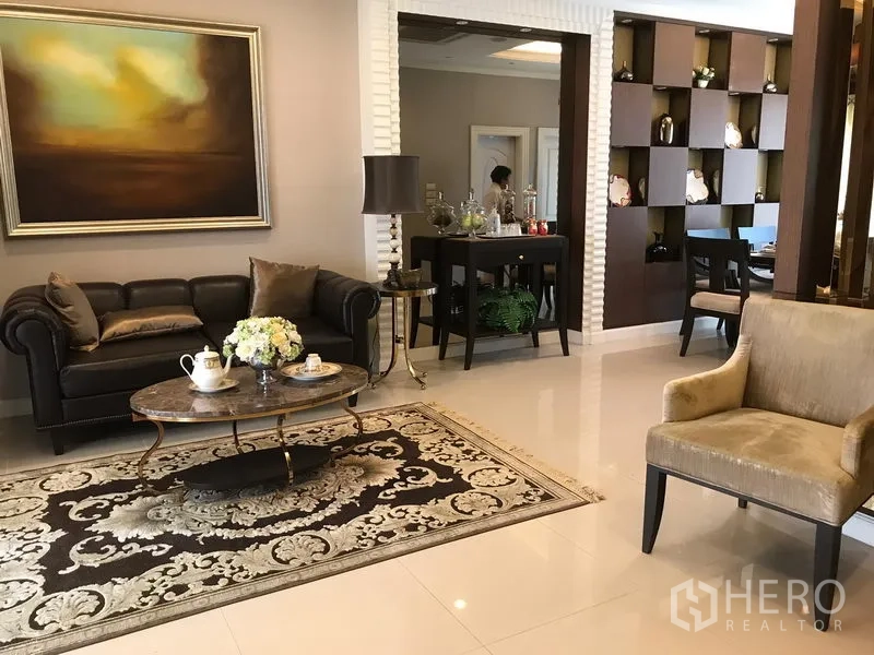 Townhouse for rent or sale in Bang Kho Laem, Bangkok - Open-plan lounge with sofa set, ornate rug and side tables, looking toward a dining space with decorative shelving.