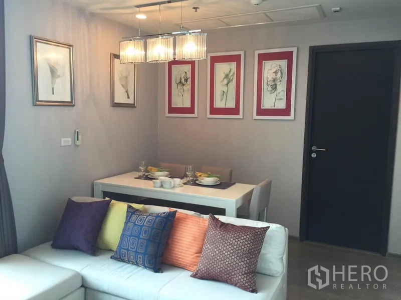 Condominium for rent in Ratchathewi, Bangkok - Living zone with sectional sofa and colorful cushions beside a bright dining table and wall art.