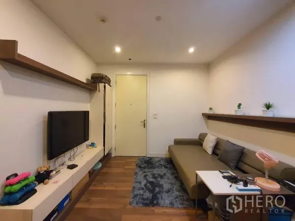 Condominium for sale in Bangkok - Cozy living room with wall-mounted TV, floating shelves, L-shaped sofa, and warm wood flooring.