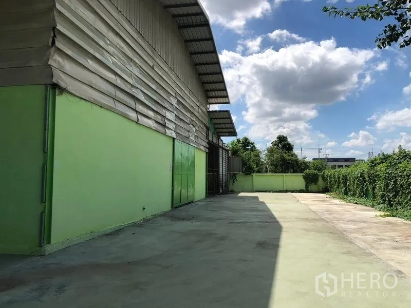 Factory for rent in Sam Khok, Pathum Thani - Factory exterior with green walls and a wide concrete yard bordered by hedges under a blue sky.