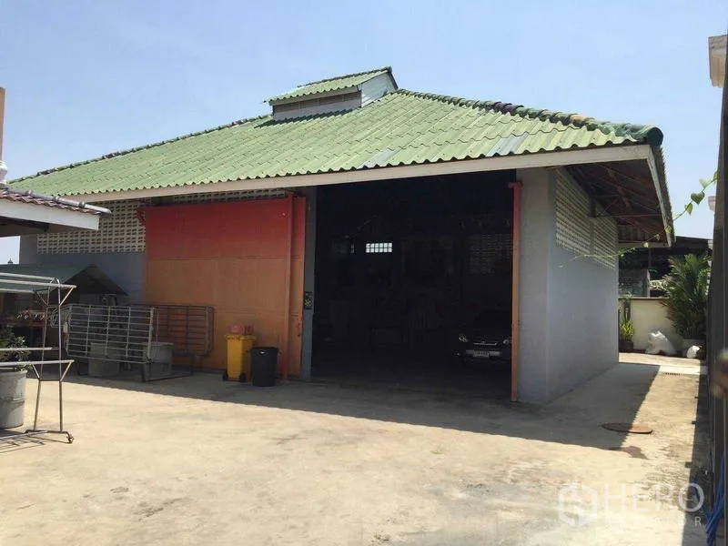 Detached House for rent or sale in Bang Bua Thong, Nonthaburi - Large detached workshop or garage with green roof and open entrance on a concrete yard.