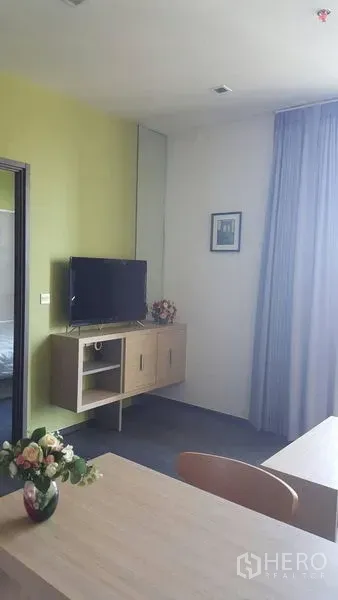 Condominium for rent in Watthana, Bangkok - Wall-mounted TV on floating cabinet beside tall curtains and dining table with a flower vase.