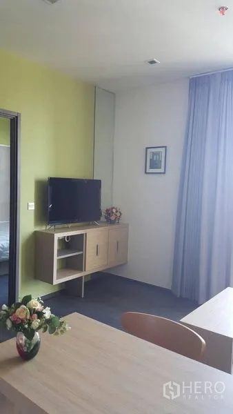 Condominium for rent in Watthana, Bangkok - Wall-mounted TV on floating cabinet beside tall curtains and dining table with a flower vase.