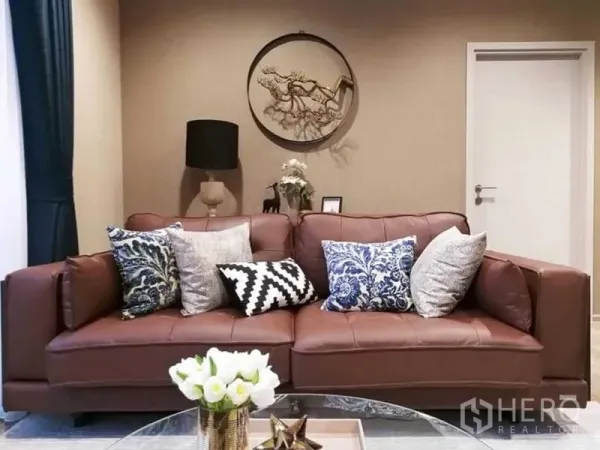 Condominium for rent in Chatuchak, Bangkok - Cozy living room with a brown leather sofa, patterned cushions, round wall art and a glass coffee table.