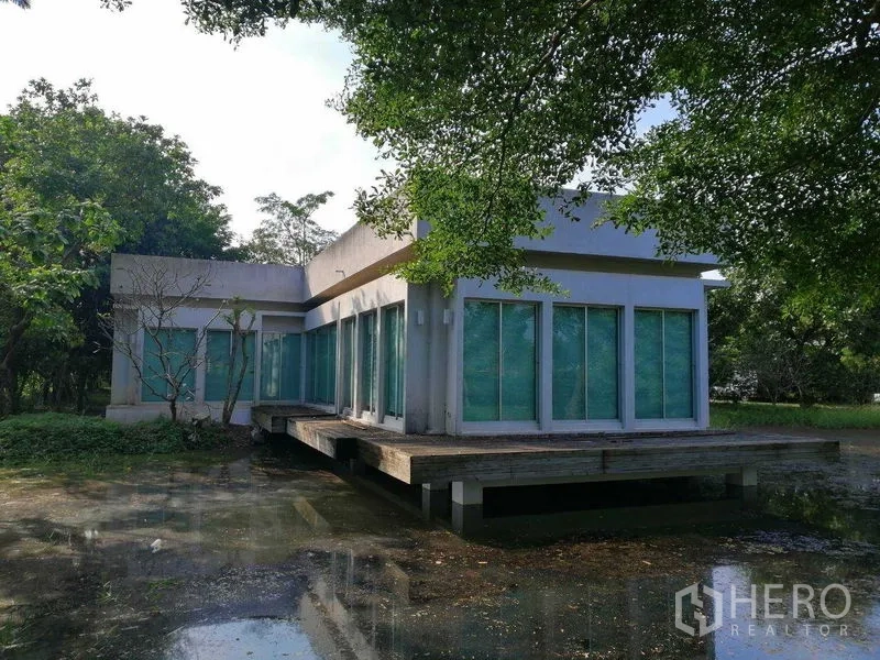 Factory for rent in Pathum Thani - Modern low-rise glass building on stilts above a pond, surrounded by mature greenery.