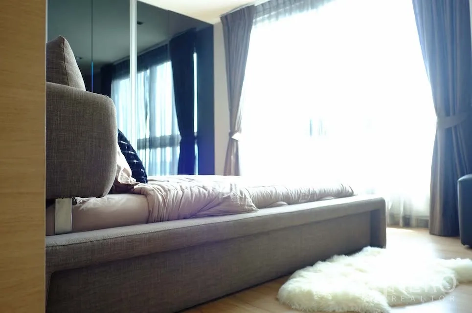 Condominium for rent or sale in Si Racha, Chon Buri - Cozy bedroom with upholstered bed, mirrored wardrobe, large curtained windows and soft rug with abundant natural light.
