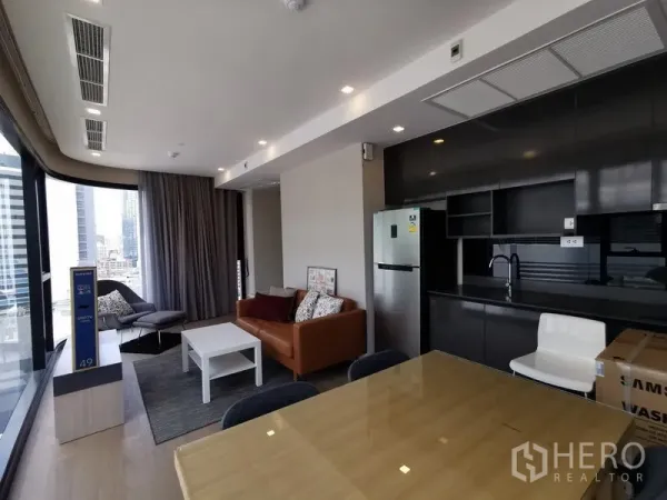 Condominium for rent or sale in Watthana, Bangkok - Open‑plan living and dining area beside full‑height windows, with a sleek black kitchen and stainless refrigerator.