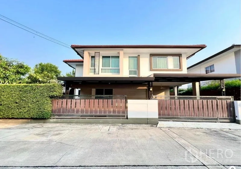 บ้าน ขายในBang Kruai, Nonthaburi - บ้านเดี่ยวสองชั้นพร้อมที่จอดรถมีหลังคาและแนวรั้วต้นไม้ บนถนนที่เงียบสงบ