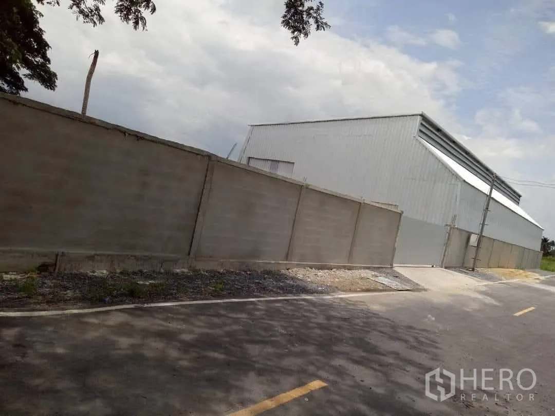 Warehouse for sale in Lam Luk Ka, Pathum Thani - Steel-clad warehouse exterior set behind a high perimeter wall along a paved access road under open sky.