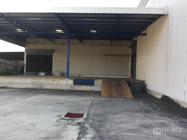 Warehouse for rent in Pak Kret, Nonthaburi - Dock-high loading area with canopy and ramp for truck access beside corrugated metal wall.