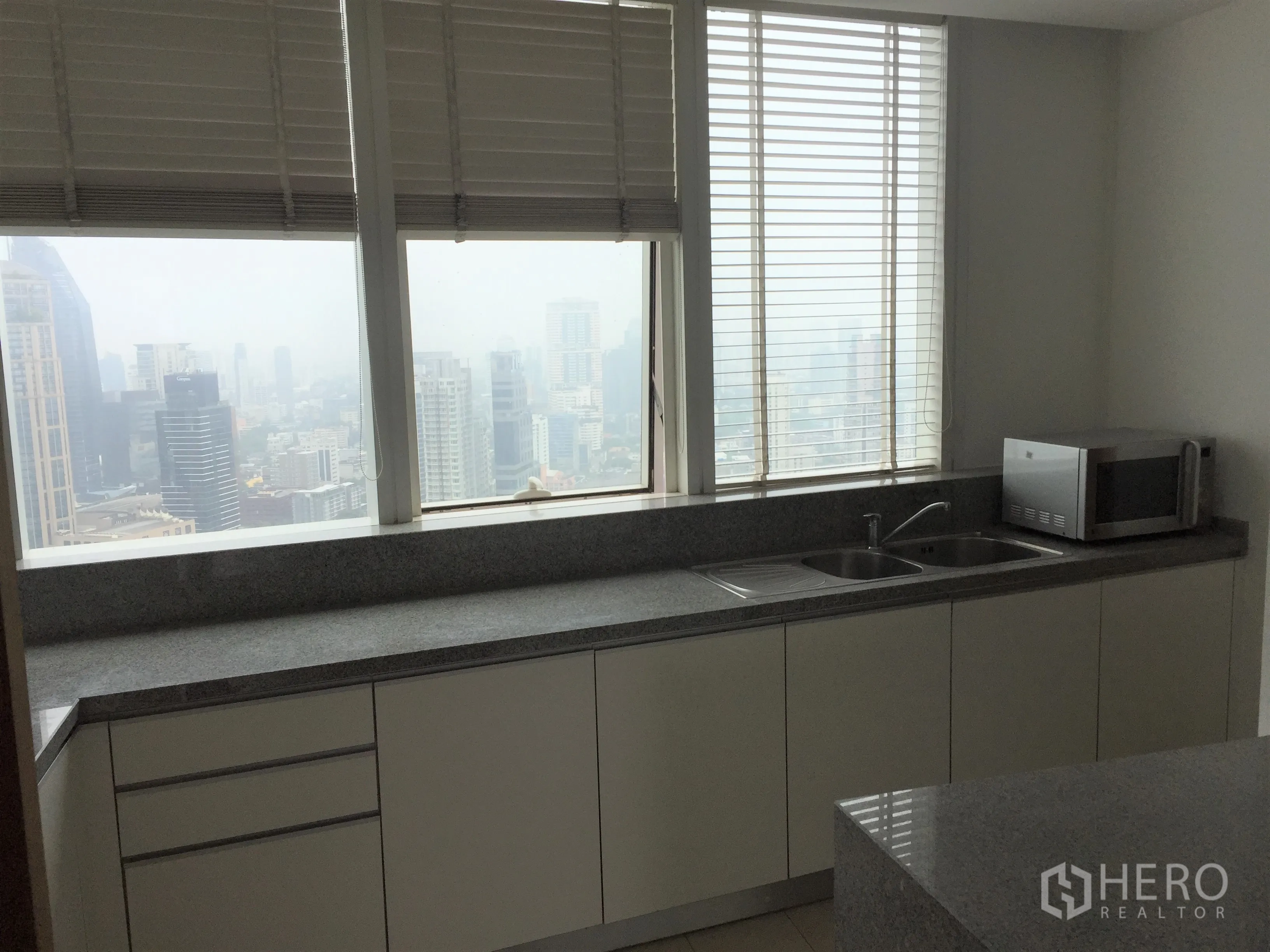 คอนโด ให้เช่าในKhlong Toei, Bangkok - ครัวมีหน้าต่าง เคาน์เตอร์หินแกรนิตยาว อ่างล้างจานคู่ ไมโครเวฟ และวิวเมืองผ่านบานมู่ลี่