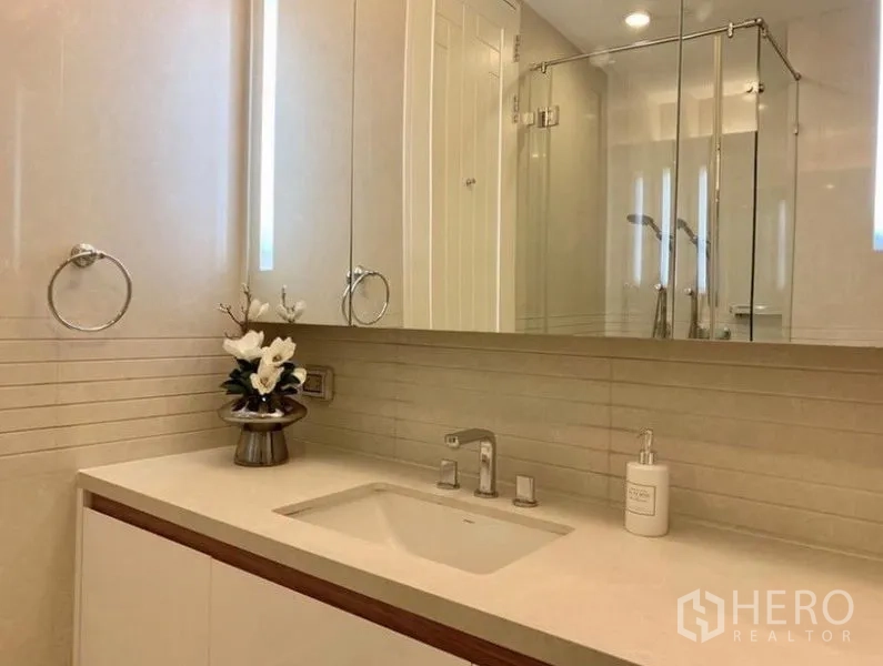 Condominium for rent in Pathum Wan, Bangkok - Modern bathroom with long vanity, under-mount sink, mirrored cabinets, chrome fixtures, and glass-enclosed shower in neutral tones.