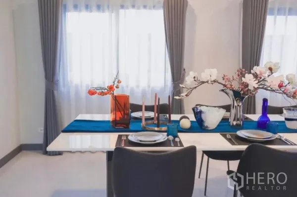 独立式住宅 出租在Bang Phli, Samut Prakan - 餐桌临双窗，灰色窗帘与花艺摆设点缀。