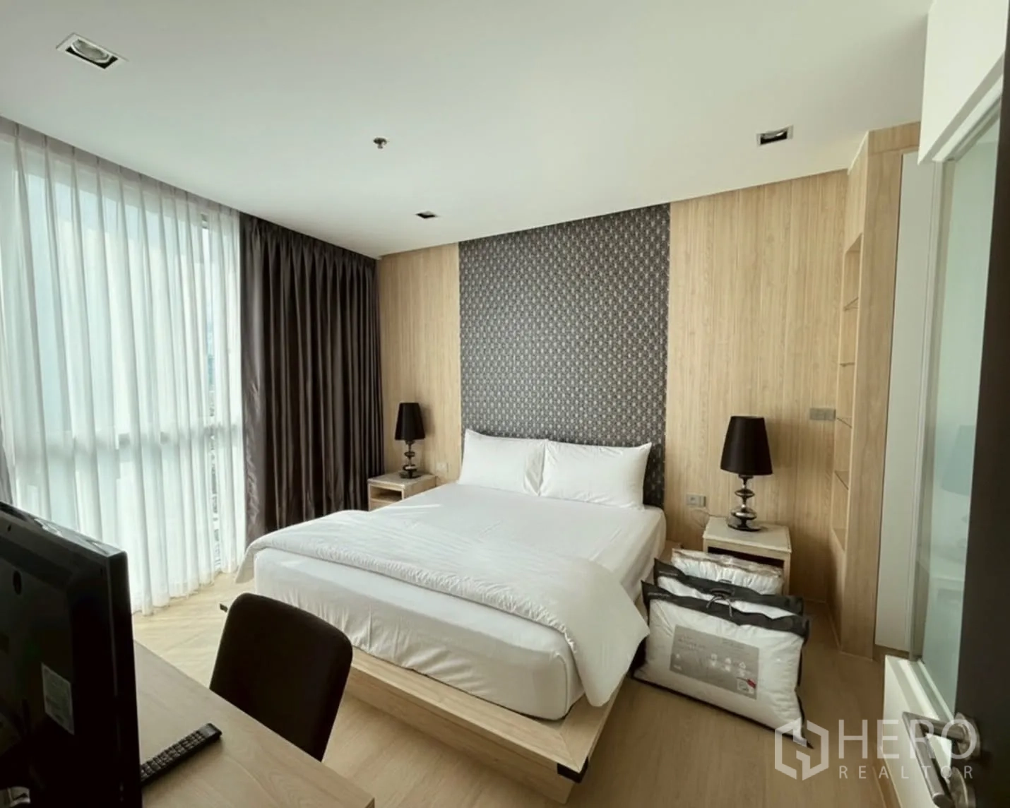 Condominium for rent or sale in Watthana, Bangkok - Bright bedroom with large bed, floor-to-ceiling windows, wood accents, and bedside lamps beside a desk.