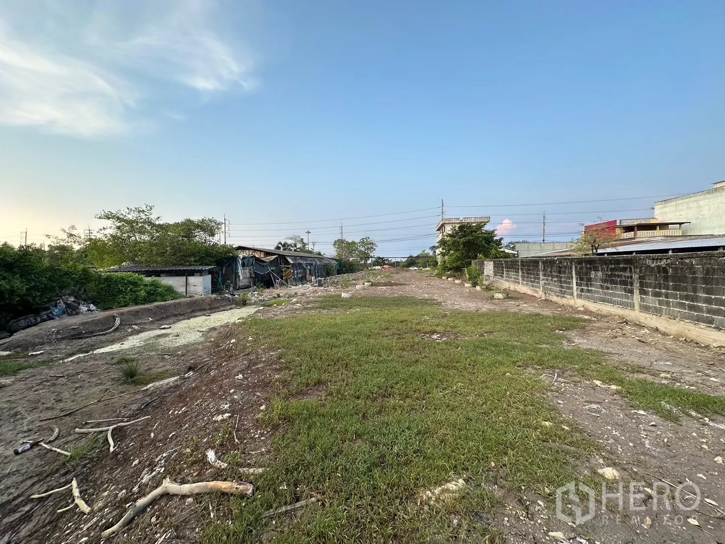 Land for sale in Mueang Samut Prakan, Samut Prakan - Cleared land plot with grass patches, boundary wall on one side, and low-rise structures nearby.
