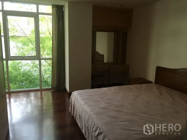 Condominium for rent or sale in Watthana, Bangkok - Primary bedroom with large window overlooking greenery, wood floors and a dressing table.