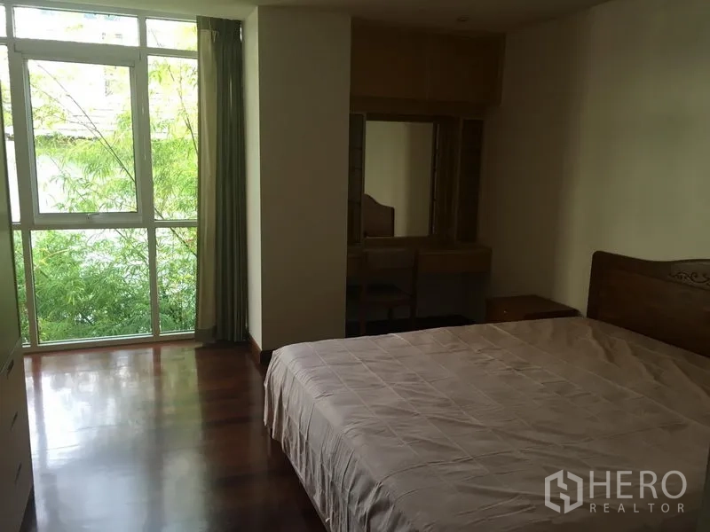 Condominium for rent or sale in Watthana, Bangkok - Primary bedroom with large window overlooking greenery, wood floors and a dressing table.