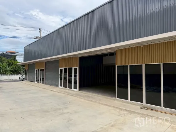 货栈 出租在Suan Luang, Bangkok - 仓库排建筑斜视图，深色波纹金属外墙，多扇玻璃入口，宽车道可供卡车通行。