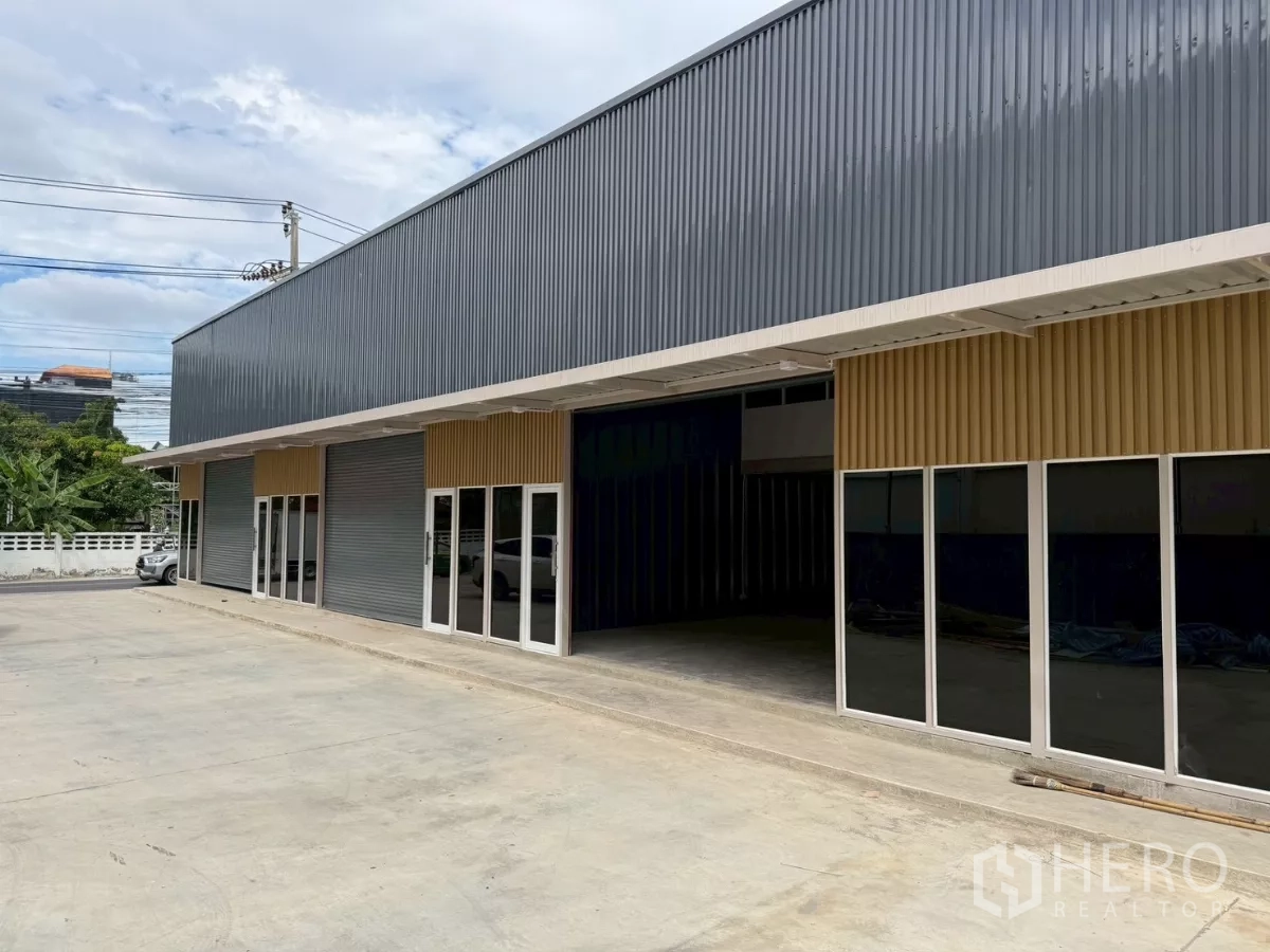 货栈 出租在Suan Luang, Bangkok - 仓库排建筑斜视图，深色波纹金属外墙，多扇玻璃入口，宽车道可供卡车通行。