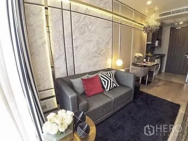 Condominium for rent or sale in Watthana, Bangkok - Cozy living area with gray sofa, accent pillows, marble wall panels, plush rug, dining nook and kitchenette.