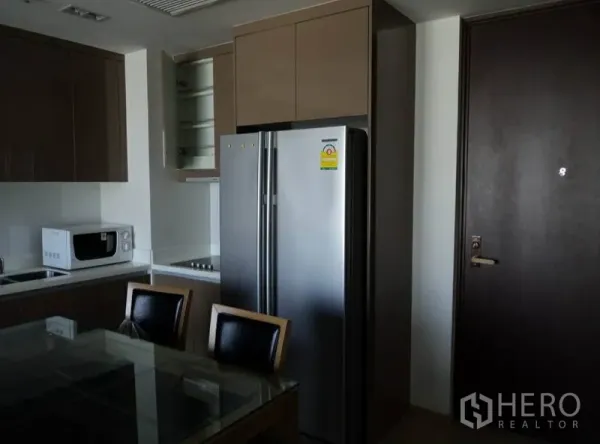 Condominium for rent in Khlong Toei, Bangkok - Compact condo kitchen with side-by-side fridge, microwave, cooktop, and a glass dining table.
