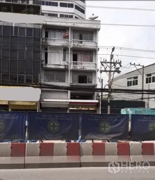 店屋 出租在Huai Khwang, Bangkok - 四层白色商住楼外立面，带阳台与一层店面，临曼谷主干道，前方有电线与施工围挡。