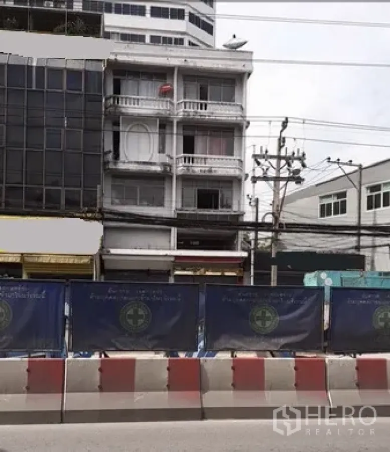 店屋 出租在Huai Khwang, Bangkok - 四层白色商住楼外立面，带阳台与一层店面，临曼谷主干道，前方有电线与施工围挡。
