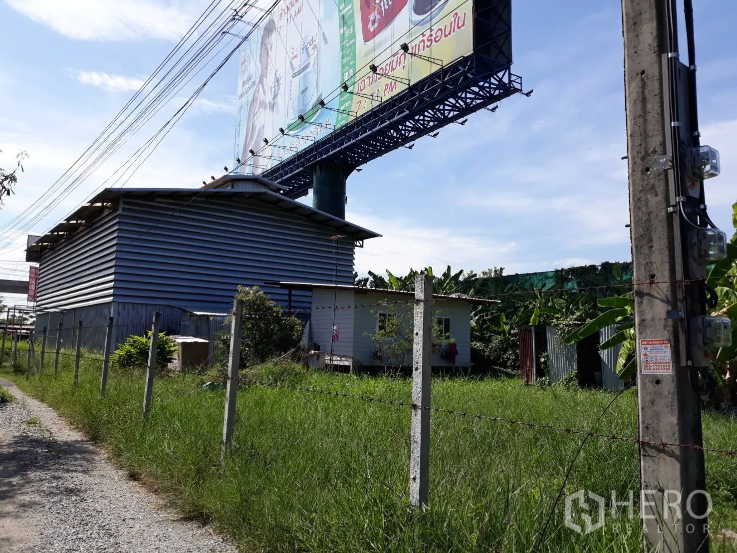 Condominium for rent in Pathum Wan, Bangkok - Side view of a corrugated metal structure and small outbuilding behind a barbed-wire fence with a large roadside billboard.
