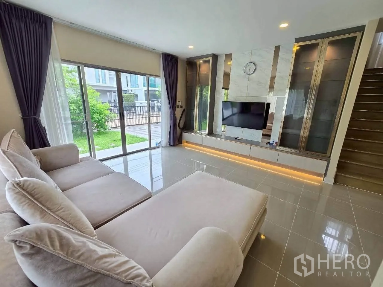 Detached House for rent or sale in Lat Krabang, Bangkok - Living room with sectional sofa, built-in TV wall, and sliding doors to garden.