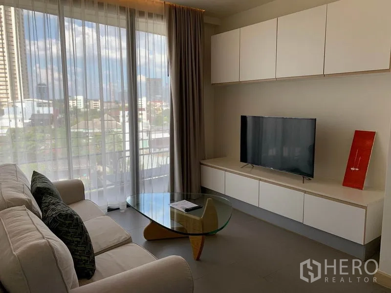 Condominium for rent in Watthana, Bangkok - Bright living room with floor-to-ceiling windows, sheer curtains, built-in TV cabinet and glass coffee table overlooking the Ekkamai skyline.