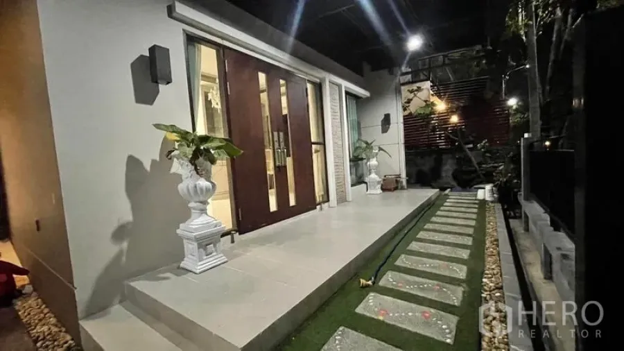 Detached House for rent or sale in Prawet, Bangkok - Night view of the front porch with double wooden doors, tiled steps, stepping‑stone path and potted plants.