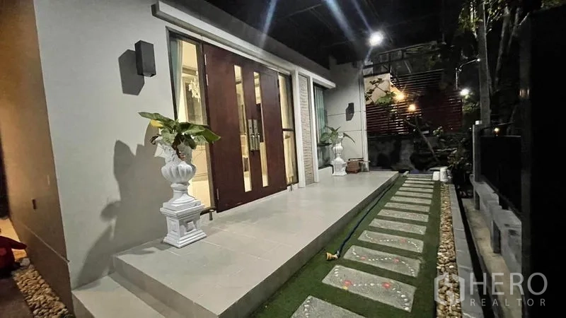 Detached House for rent or sale in Prawet, Bangkok - Night view of the front porch with double wooden doors, tiled steps, stepping‑stone path and potted plants.