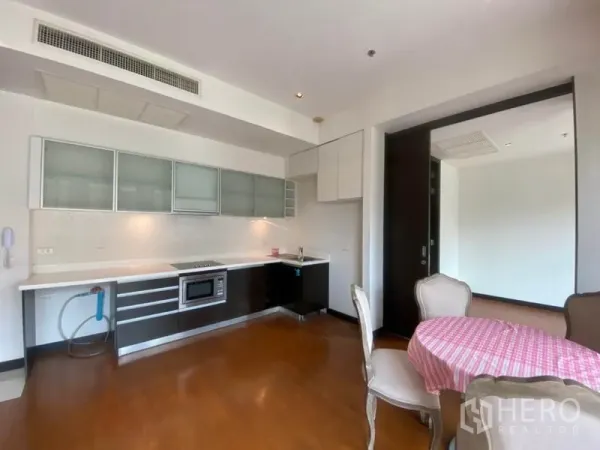 Condominium for rent in Yan Nawa, Bangkok - Modern kitchenette with white countertop, glass-front cabinets, electric cooktop and built-in oven beside a small dining table.