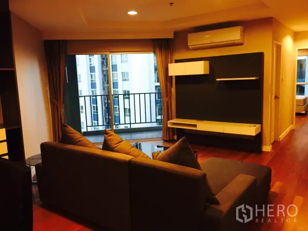 Condominium for rent in Huai Khwang, Bangkok - Cozy living room with sectional sofa, wood floors, TV wall and balcony overlooking city buildings.
