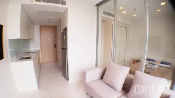 Condominium for rent in Watthana, Bangkok - Compact open kitchen with built-in cabinets and fridge beside a sofa and glass-walled bedroom in the 1-bed condo.