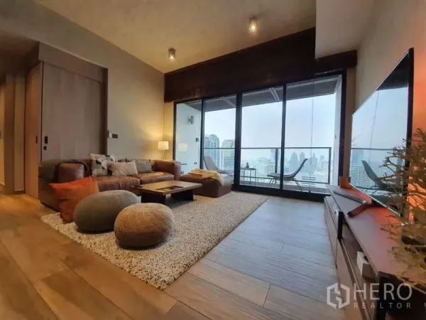 Condominium for rent or sale in Watthana, Bangkok - Warm-toned living room with leather sofa, rug, and floor-to-ceiling windows opening to a balcony with Bangkok skyline views.