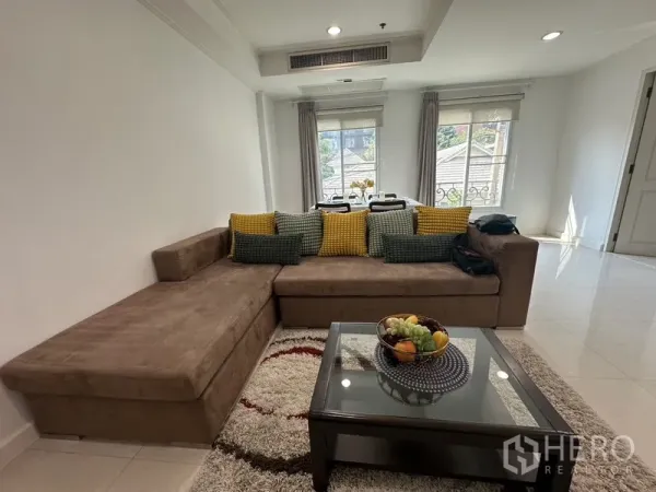 公寓 出租 - 明亮客厅，L形沙发、地毯与玻璃茶几，后方为窗边餐区