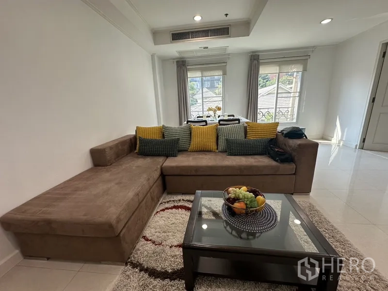 公寓 出租 - 明亮客厅，L形沙发、地毯与玻璃茶几，后方为窗边餐区