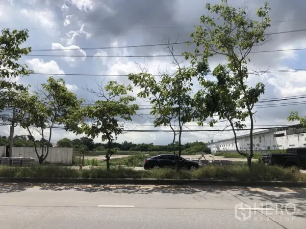 Land for sale in Mueang Pathum Thani, Pathum Thani - Roadside view of a large vacant land plot in Mueang Pathum Thani with trees, power lines, and nearby warehouse buildings.