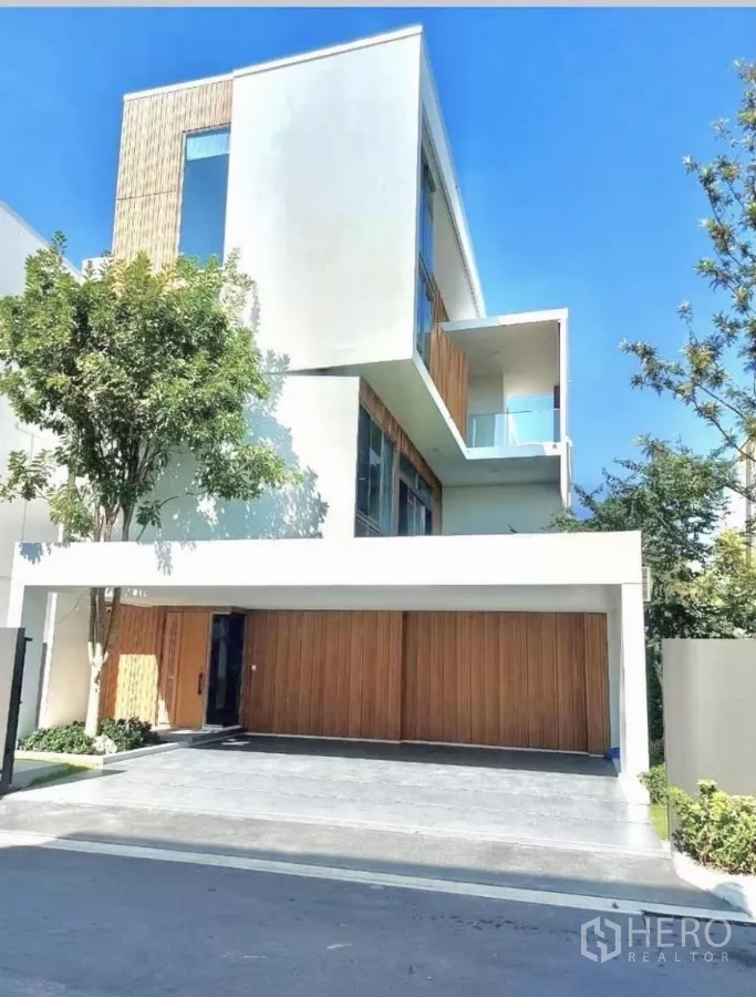 Detached House for rent in Saphan Sung, Bangkok - Modern three-storey detached house facade with wood garage doors, large windows and a cantilevered balcony under a clear sky.