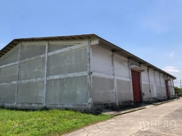 คลังสินค้า ให้เช่าในLat Krabang, Bangkok - ภาพมุมกว้างด้านนอกโกดังคอนกรีตชั้นเดียว ประตูสไลด์สีแดง หลังคาเมทัลชีท และทางเข้าเทคอนกรีตใต้ท้องฟ้าโปร่ง