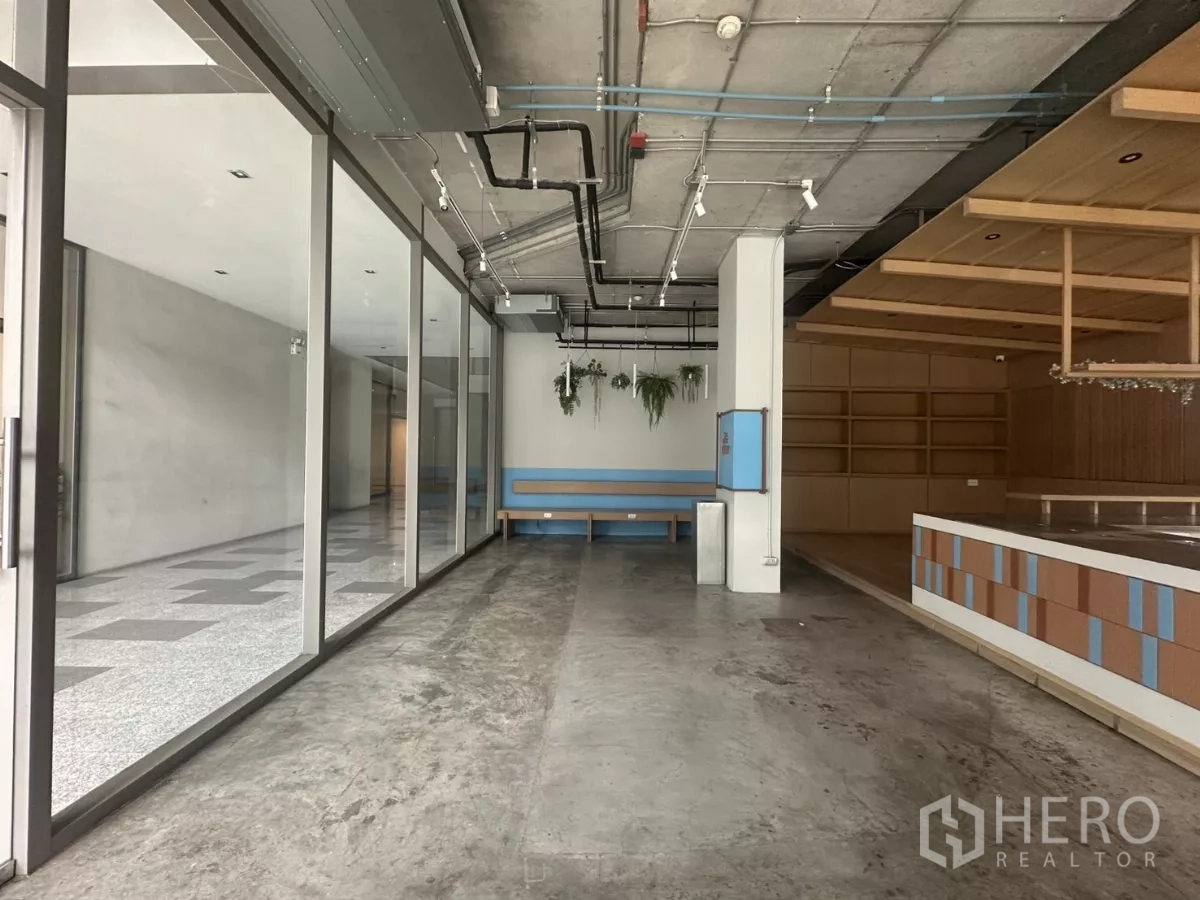 พื้นที่ขายปลีก ให้เช่า - มุมมองยาวตามหน้าร้านกระจก เห็นฝ้าเพดานเปลือย ไฟราง และเคาน์เตอร์ยกระดับ